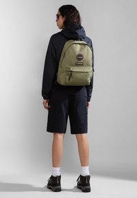 Zaino verde oliva con spallacci imbottiti, tasca frontale e patch con logo. Indossato sopra una giacca nera e pantaloni corti, su uno sfondo neutro.