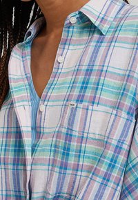 Camicia a quadri a maniche corte, caratterizzata da strisce blu, verdi e viola su uno sfondo bianco, include una tasca sul petto e chiusura con bottoni.