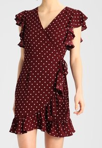 Robe portefeuille bordeaux à pois blancs, avec des manches courtes à volants, un ourlet festonné et un détail de nœud sur le côté sur le torse d'un mannequin.