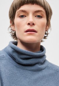 Modra pletena pulover s turtleneck izjemno mehke teksture in zloženim ovratnikom, ki ima subtilen vzorec in sproščen kroj. Vidne srebrne obročeve uhane.