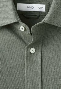 Polo de tela verde oliva texturizada que presenta un cuello clásico y dos botones blancos en el escote. La etiqueta dice "MNG EU-USA M MEX M."