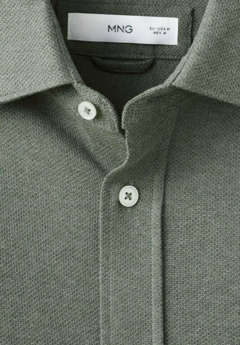 Polo-skjorte i tekstureret olivengrønt stof med en klassisk krave og to hvide knapper ved halsen. Etiketten siger "MNG EU-USA M MEX M."