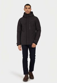 Veste imperméable noire avec capuche, fermeture éclair à l'avant, poche poitrine et deux poches latérales inférieures. Portée avec un jean foncé et des bottes marron.