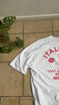Hvid T-shirt med rød italiensk inspireret print, som viser en bøjet arm, tekst og ternet mønster på en tandfarvet flisegulv nær en grøn potteplante.