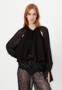 Zwarte, doorschijnende blouse met een geplooide halslijn, verlengde mouwen en uitsnijdingen op de schouders. Gepaard met zwarte hoge taille shorts met pailletten.