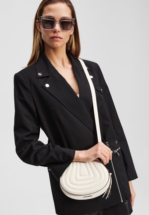 Geantă de umăr albă, cu un design curbat, purtată pe umăr. Model îmbrăcat cu un sacou negru cu accente argintii și ochelari de soare supradimensionați.