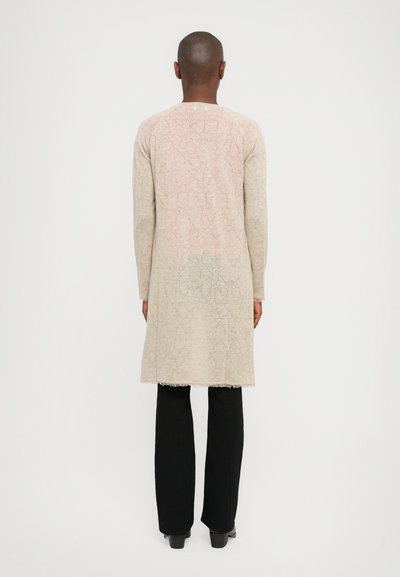 Long, knitted cardigan in light beige with a gradient effect from pink to green. Features a geometric pattern and frayed hem. Worn with black pants.