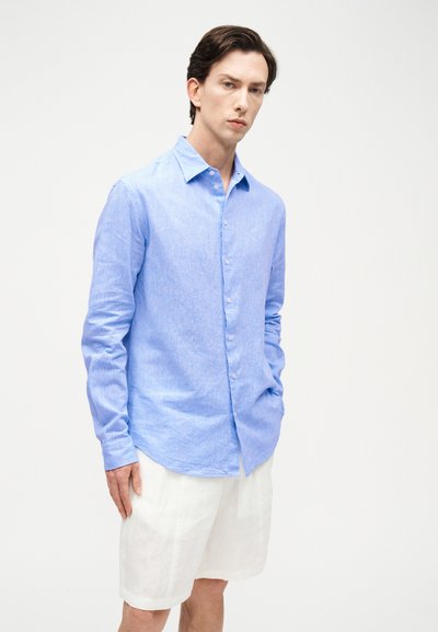 Νεαρός άνδρας που φοράει ανοιχτό γαλάζιο πουκάμισο με μακριά μανίκια και λευκά σορτς μέχρι το γόνατο, σε απλό λευκό φόντο.