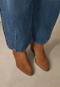 Bruine suède enkellaarsjes met puntige neuzen, gestapelde hakken en subtiele stiksels, gecombineerd met blauwe denim wijd uitlopende jeans.