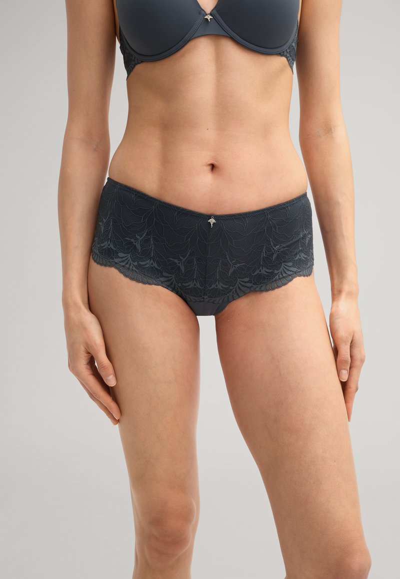 Culotte hipster en dentelle gris foncé avec un bord festonné, un motif floral et un accent en métal décoratif au centre devant. Tissu doux au toucher.