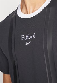 Zwarte T-shirt met witte ronde halslijn, voorzien van de tekst "Fútbol" en het Nike-logo in witte borduursel. Verticale zwarte strepen voegen textuur toe.
