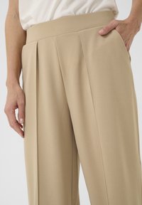 Beige tailored trousers with a high waistband, featuring sharp pleats and side pockets. Smooth texture, structured fit, and minimalistic design.
