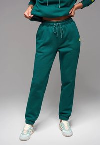 Pantalones de chándal verdes hechos de una tela suave, con una cinturilla elástica con cordón, tobillos ajustados y un pequeño acento de logo.