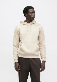 Könnyű, puha anyagból készült bézs kapucnis pulóver, elülső zsebbel, húzózsinórokkal és a mellkason kis fehér logóval. Sötét színű melegítőnadrággal párosítva.