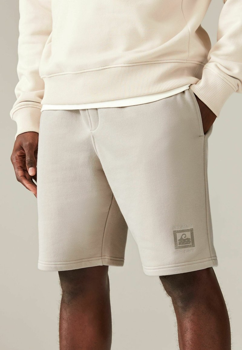 Mand iført beige shorts med "Miami Coast" logo på venstre ben og en cremefarvet sweater, hænderne i lommerne, stående mod en neutral baggrund.