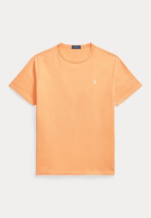 Απλό πορτοκαλί κοντομάνικο T-shirt με στρογγυλή λαιμόκοψη, με μικρό λευκό κεντημένο λογότυπο στο αριστερό μέρος του στήθους και μια μπλε ετικέτα μέσα στο γιακά.
