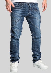 Denim jeans in dark blue with a fitted design, featuring unique diagonal seams, multiple pockets, and light stitching, paired with white sneakers.