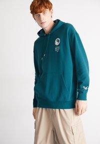 Teal mikina s kapucňou a predným vreckom, zlaté aglety na šnúrkach, grafika Yin-Yang na hrudi a biele logo na rukáve.