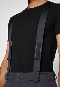 Camisa preta de mangas curtas combinada com suspensórios cinzentos com um acabamento texturizado e detalhes em Velcro ajustáveis nas tiras.