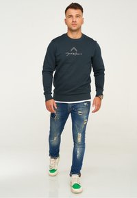 Dark teal sweatshirt with a round neckline and logo on the chest; paired with distressed blue jeans and white sneakers with green accents.