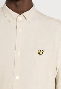 Beige napitettava paita, jossa on kaulus, vasemmassa rinnassa tasku ja keltainen brodeerattu logo. Kangas on teksturoitu.