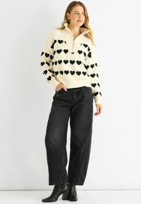 Cream sweater with black heart pattern, quarter-zip collar, paired with high-waisted black jeans and black heeled ankle boots.