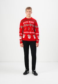 Rode gebreide trui met feestelijke patronen, waaronder de kerstman, snoepstokken en sneeuwvlokken, met de tekst "BLISSFUL HOLIDAYS" in het wit. Zwarte broek en schoenen.