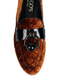 Loafer w kolorze rdzy z weluru z okrągłym wytłaczanym wzorem, czarny lakierowany skórzany pasek oraz metalowy pierścień z dużą czarną perłą jako ozdobą.