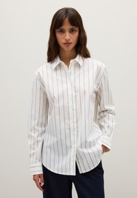 Mujer vistiendo una camisa blanca de botones con finas rayas verticales negras y pantalones oscuros, de pie con una mano en el bolsillo frente a un fondo liso.