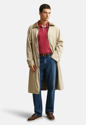 Mann trägt beigen Trenchcoat über rotem Poloshirt, blaue Jeans, braunen Gürtel und braune Freizeitschuhe, steht mit einer Hand in der Hosentasche.