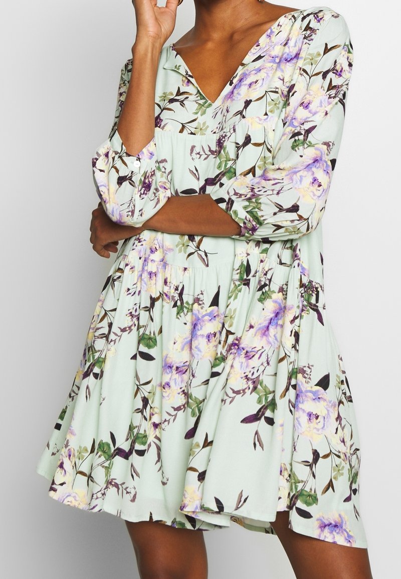 Robe fleurie en tissu vert clair avec un décolleté en V, des manches 3/4 et une taille froncée. Présente des motifs floraux violets et jaunes.