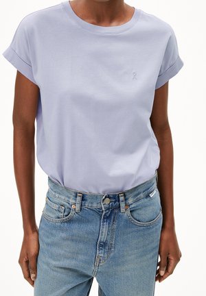 Osoba ubrana w jasnofioletową koszulę z krótkim rękawem z podwiniętymi mankietami, włożoną do wysokiej talii niebieskich dżinsów, stojąca na białym tle.