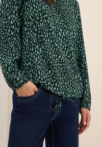 Grün gemusterte Bluse mit einem Knoten in der Taille, langärmlig und locker geschnitten, kombiniert mit dunkelblauen Jeans.