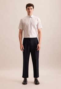 Camicia a maniche corte in gingham marrone chiaro con bottoni; abbinata a pantaloni blu a pieghe e scarpe nere con lacci, in piedi su uno sfondo neutro.