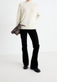 Suéter de punto crema con patrón de ondas texturizado, combinado con pantalones acampanados negros y botas negras gruesas. Sosteniendo una bolsa gris oscuro.