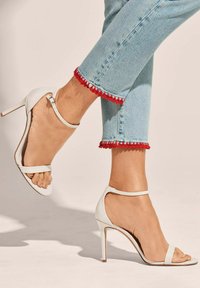 Weiße High Heel Sandalen mit einem offenen Zehen-Design, Knöchelriemen und glattem Leder. Kombiniert mit hellblauen Jeans, die mit roten dekorativen Nähten versehen sind.