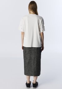 Weißes, kurzärmliges Oberteil mit Knopfdetails, kombiniert mit einem langen, grauen Tweed-Rock mit Seitentaschen und einem Schlitz hinten. Schwarze, strukturierte Schuhe.