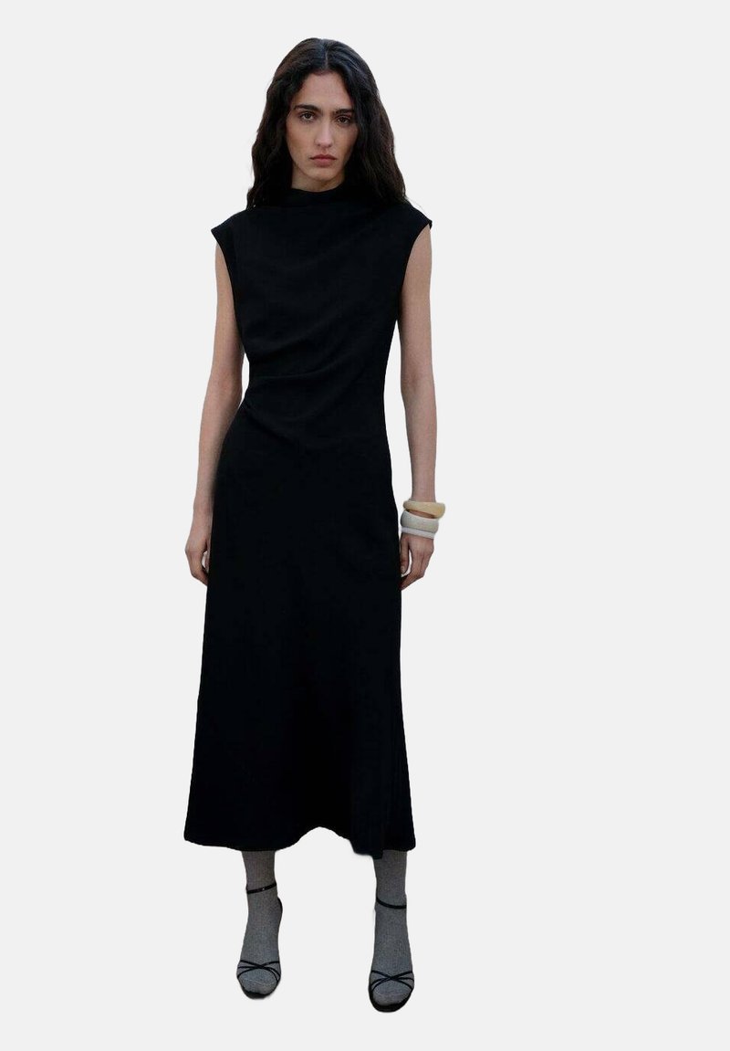 Rochie neagră fără mâneci, cu guler înalt, bust ajustat și fustă midi fluidă; țesătură netedă; completată cu brățări și șosete neutre.