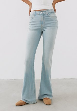 Personne portant un jean flare bleu clair, un haut blanc et des chaussures plates marron, debout sur un sol gris uni contre un mur blanc.