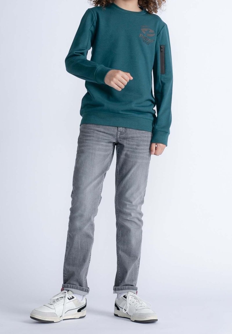 Enfant portant un sweat-shirt turquoise, un jean gris et des baskets blanches, debout devant un fond clair.