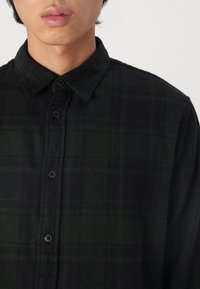 Black and green plaid button-up shirt with a collar, featuring a textured fabric and black buttons along the front.