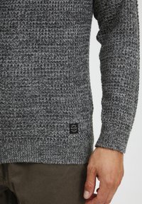 Grå strikket sweater med et tekstureret mønster, med en lille sort logo patch på kanten og lange ærmer.
