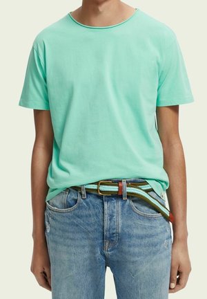 T-shirt en coton turquoise avec un col rond, associé à un jean en denim bleu clair. La tenue est complétée par une ceinture tissée multicolore avec une boucle en métal.