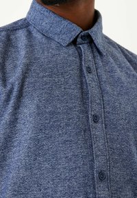 Modrá texturovaná košile s designem na zapínání na knoflíky, standardním límcem a tmavě modrými knoflíky podél légy.