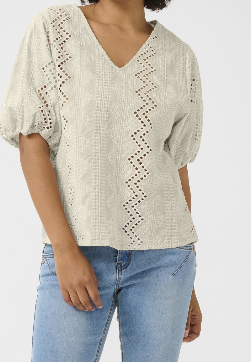 Femme portant un chemisier crème texturé avec des motifs découpés en zigzag et des manches courtes bouffantes, associé à un jean bleu clair.