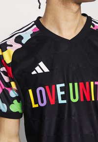 Czarna koszulka sportowa z kolorowym, abstrakcyjnym wzorem na rękawach. Z przodu widoczny jest wyrazisty napis „LOVE UNITE” w wielu kolorach.