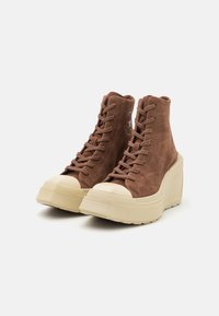 Adidași înalți din suede maro cu design cu șireturi, vârfuri din cauciuc și talpă groasă de culoare crem. Prezintă un logo pe lateral.