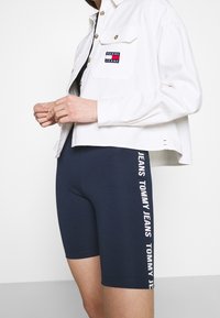 Vit kortärmad jacka med knäppning och bröstlogotyp; marinblå cykelshorts med "TOMMY JEANS" tryckt vertikalt i vitt längs sidan.
