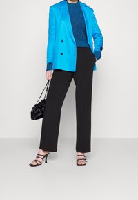 Modèle portant un blazer bleu vif et un pantalon sombre, tenant un sac noir. Elle porte des talons noirs à lanières, se tenant devant un fond clair.