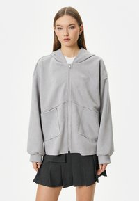 Grå zip-up hoodie lavet af blødt stof, med hætte, to frontlommer og løse ærmer. Bæres over en hvid skjorte og sort nederdel.